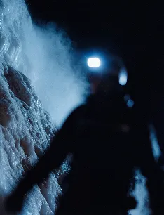 Silhouette of a person wearing a headlamp, standing near an ice wall at night.