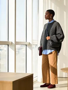 Woman in gray knit cardigan, blue shirt, and tan pants standing by a large window with sunlight streaming in.